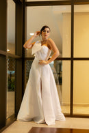 Na imagem, uma mulher veste o elegante vestido de noiva "Pilar" da coleção Barcelona da Luuly Noivas. O vestido é confeccionado em tecido Zibeline branco, com um decote coração e uma única alça larga no ombro esquerdo, adornada com um laço estruturado. A silhueta é longa e fluida, com uma fenda frontal elegante. A mulher está de pé em frente a uma janela, destacando a sofisticação e o design moderno do vestido.  Porque o amor não pode esperar.