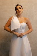 Na imagem, uma mulher está usando o vestido de noiva "Pilar" da coleção Barcelona da Luuly Noivas. O vestido é confeccionado em tecido Zibeline branco, destacando um decote coração e uma única alça larga no ombro esquerdo, adornada com um laço estruturado. A silhueta é longa e fluida, com um design elegante e moderno. A mulher está de pé contra uma parede de fundo neutro, com as mãos cruzadas na frente, destacando a sofisticação e o detalhe do vestido.