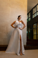 Na foto, vemos uma mulher vestindo o elegante vestido de noiva "Pilar" da coleção Barcelona da Luuly Noivas. O vestido, feito de tecido Zibeline branco, possui um decote coração e uma única alça larga no ombro esquerdo, adornada com um laço estruturado. A silhueta é longa e fluida, com uma fenda frontal que adiciona um toque sofisticado e moderno ao visual. A mulher está de pé, olhando ligeiramente para o lado, destacando a beleza e os detalhes únicos do vestido.