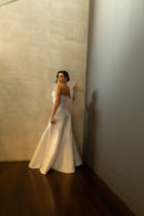 O vestido de noiva "Pilar" da coleção Barcelona da Luuly Noivas é um modelo sofisticado e moderno. Confeccionado em tecido Zibeline branco, este vestido longo possui uma silhueta elegante com uma fenda frontal. O design destaca-se por um decote coração e uma única alça larga no ombro esquerdo, adornada com um laço estruturado. Ideal para noivas que buscam um visual único e refinado no grande dia.